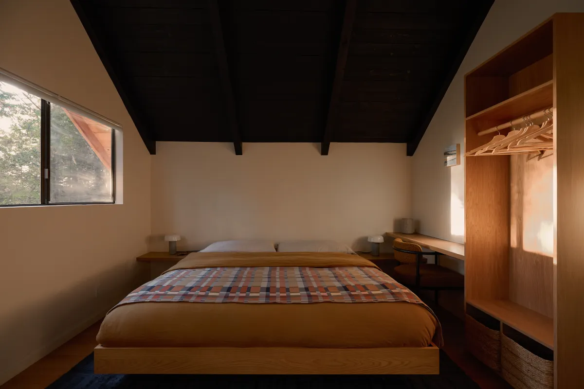 A Timber Ridge bedroom at Sea Ranch: a low oak platform bed with a mustard duvet and a checked throw, a built-in oak closet, under a dark A-frame ceiling.