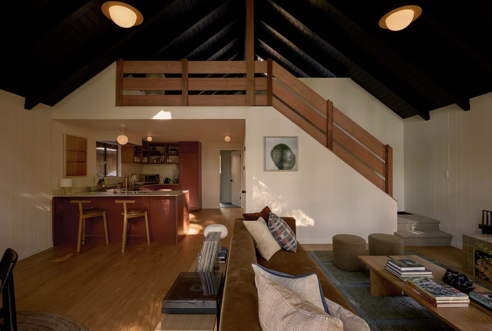 The loft and kitchen after the remodel: oxblood cabinetry, a new staircase with redwood railing, white oak floors under a blackened vaulted ceiling.